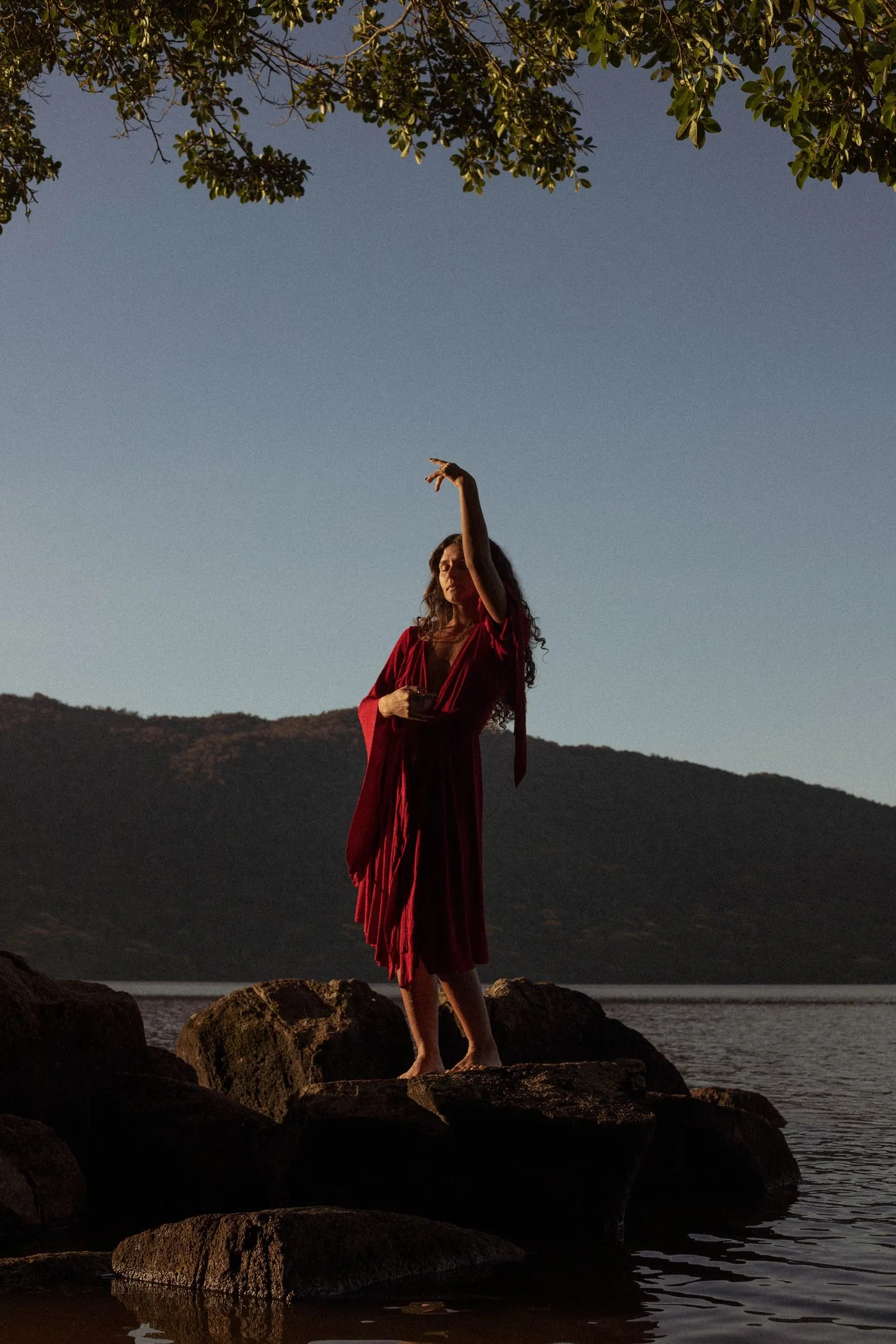 Kalina Cunha de vestido vermelho em pé sobre as pedras à beira da água, ao entardecer
