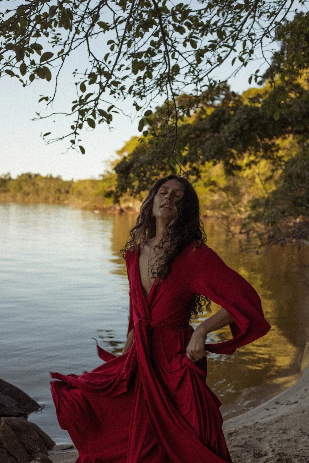 Mulher de vestido vermelho à beira da água, olhos fechados, em entrega