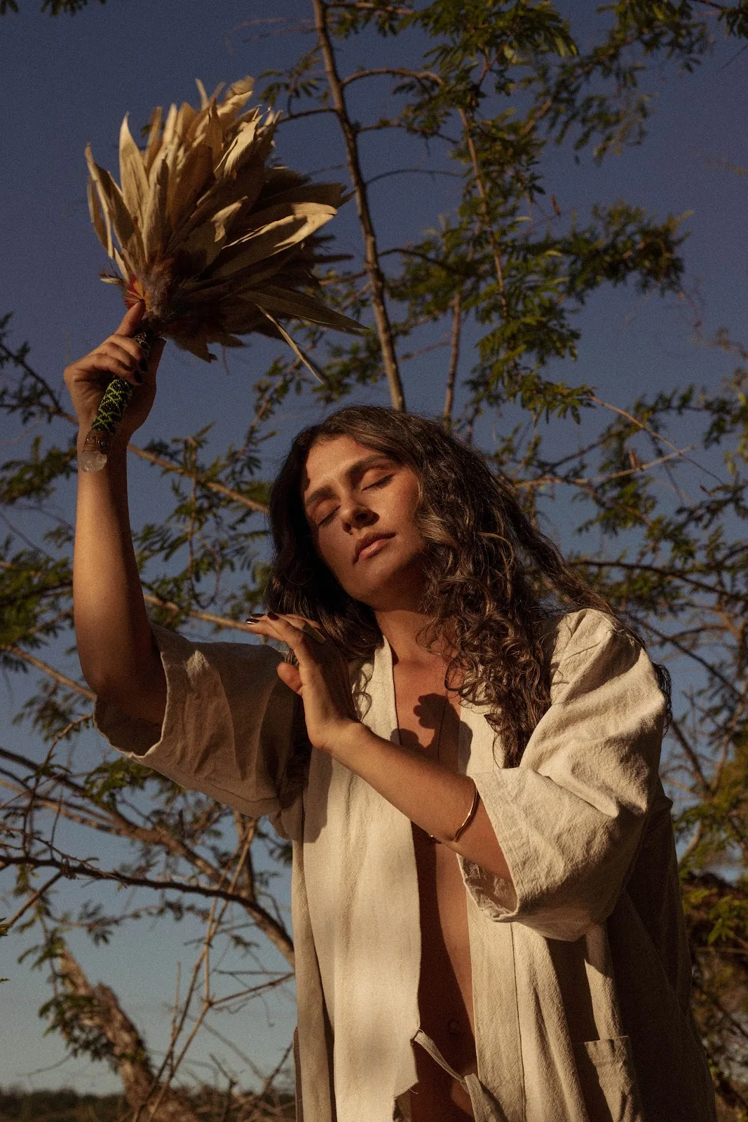 Mulher de braços erguidos segurando um leque ritual contra o céu azul profundo